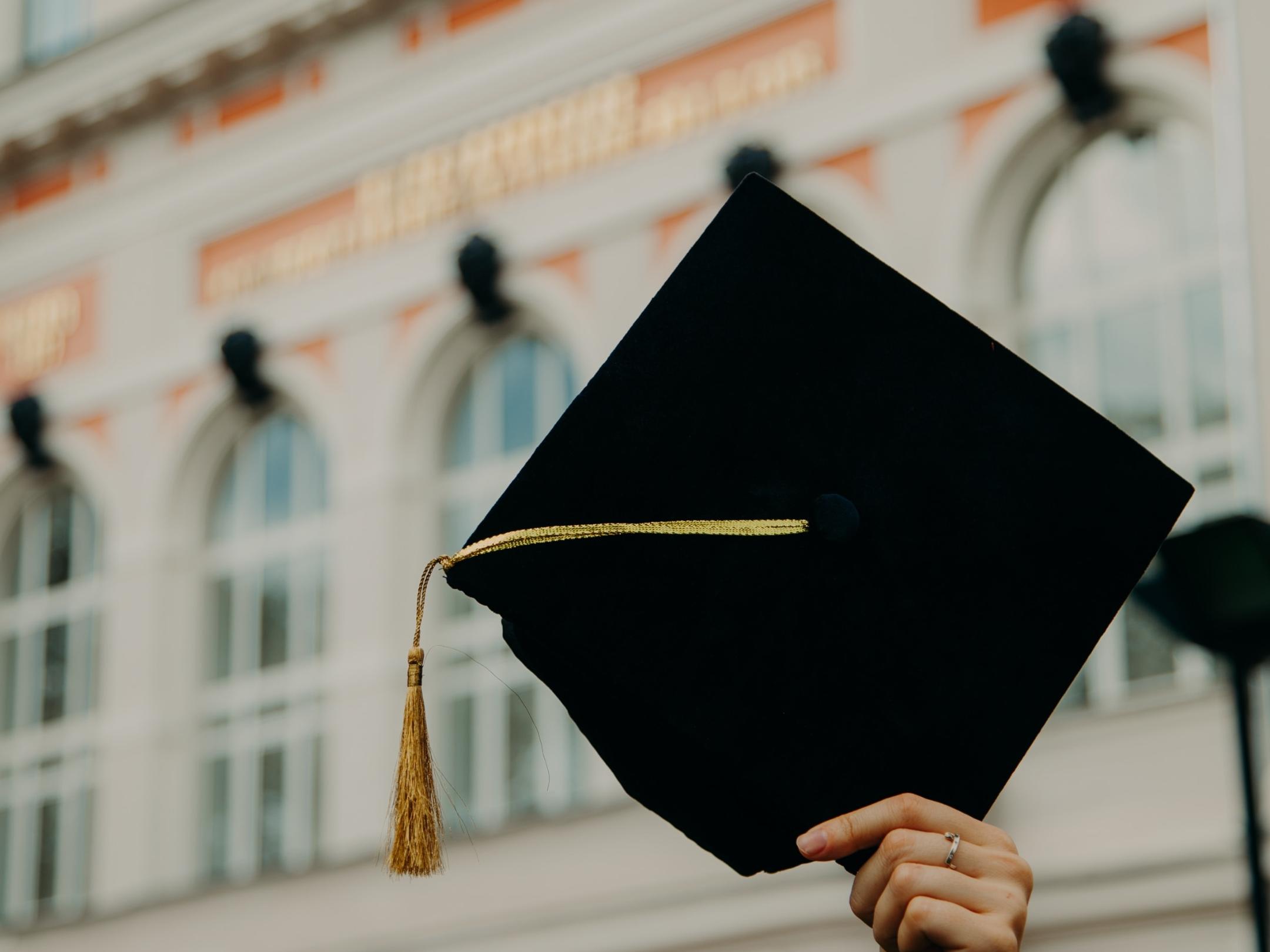 Eine Person steht vor der Universität und hält einen Doktorhut in die Höhe.