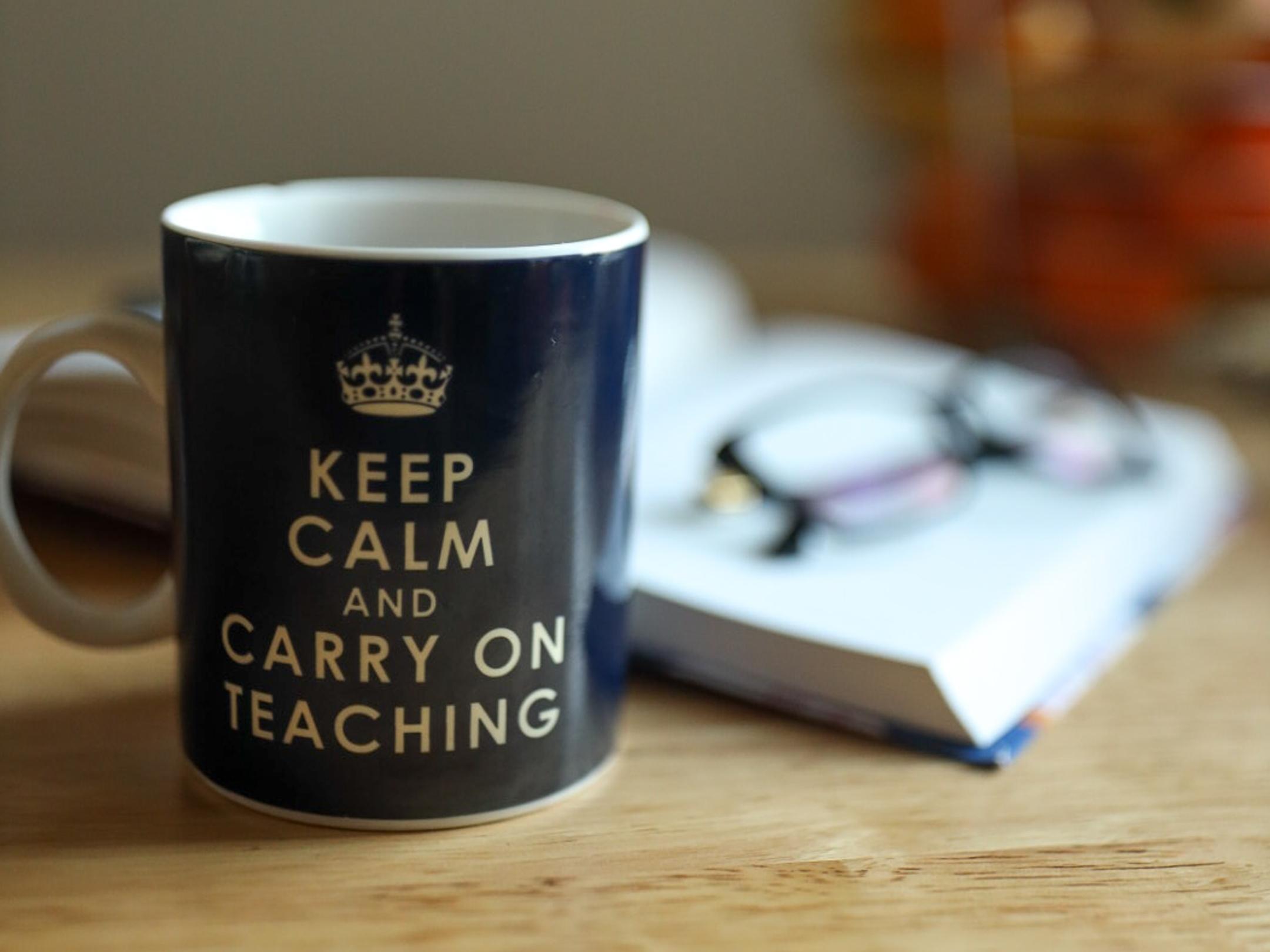 Auf einem Tisch steht eine Tasse mit der Aufschrift Keep calm and carry on teaching; im Hintergrund ein Buch mit Brille darauf und ein Korb mit Äpfeln
