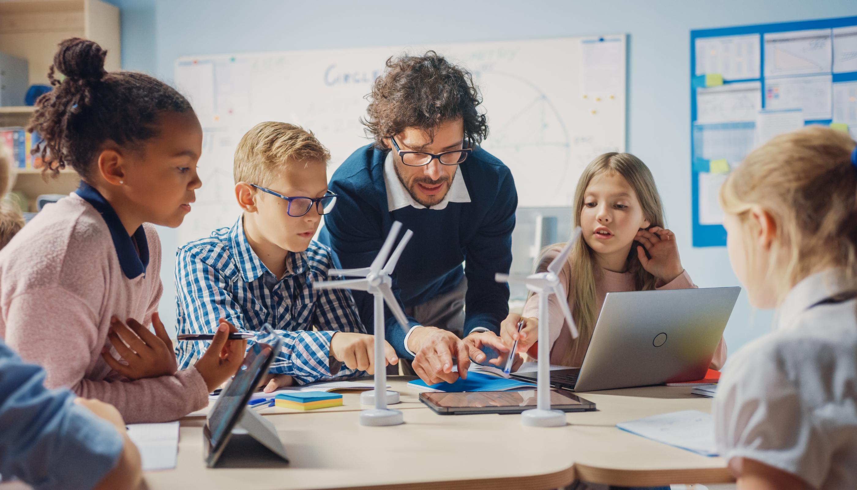 Man sieht Schüler*innen an einem Tisch mit Tablet, Laptop und Miniaturwindrädern. Ein Lehrer beugt sich zur Gruppe, um etwas zu erklären