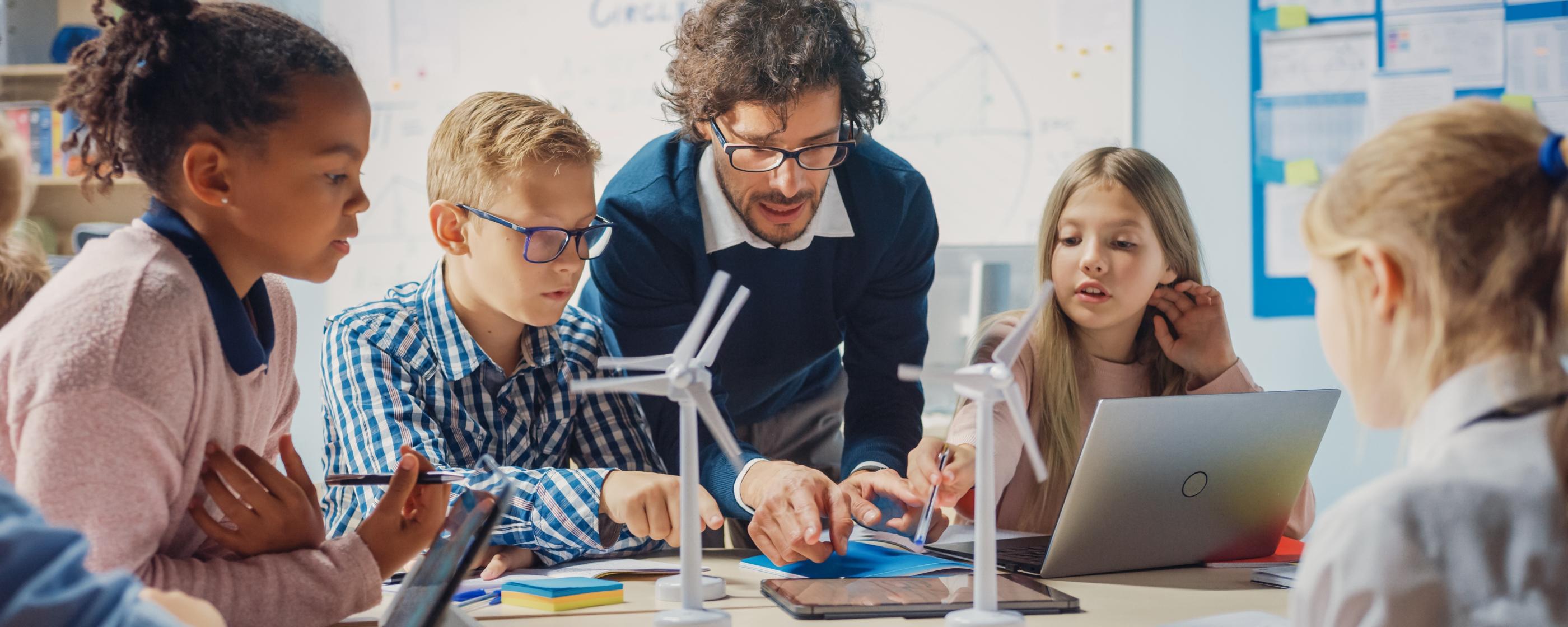 Man sieht Schüler*innen an einem Tisch mit Tablet, Laptop und Miniaturwindrädern. Ein Lehrer beugt sich zur Gruppe, um etwas zu erklären