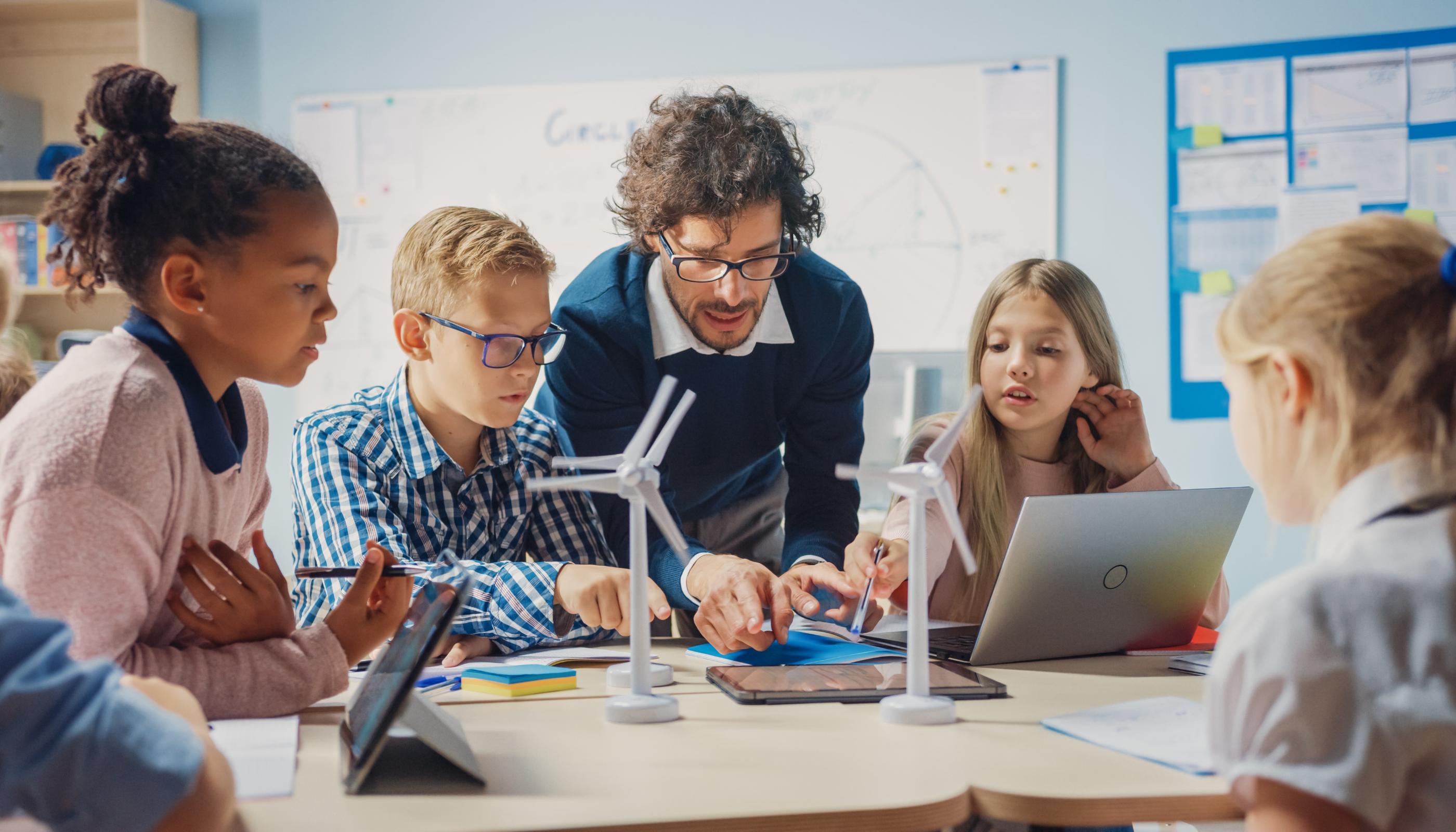 Man sieht Schüler*innen an einem Tisch mit Tablet, Laptop und Miniaturwindrädern. Ein Lehrer beugt sich zur Gruppe, um etwas zu erklären