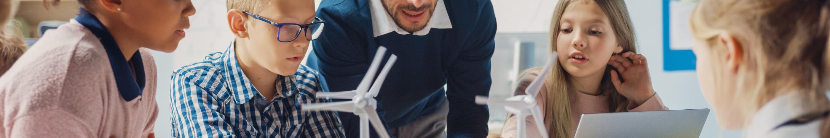 Man sieht Schüler*innen an einem Tisch mit Tablet, Laptop und Miniaturwindrädern. Ein Lehrer beugt sich zur Gruppe, um etwas zu erklären