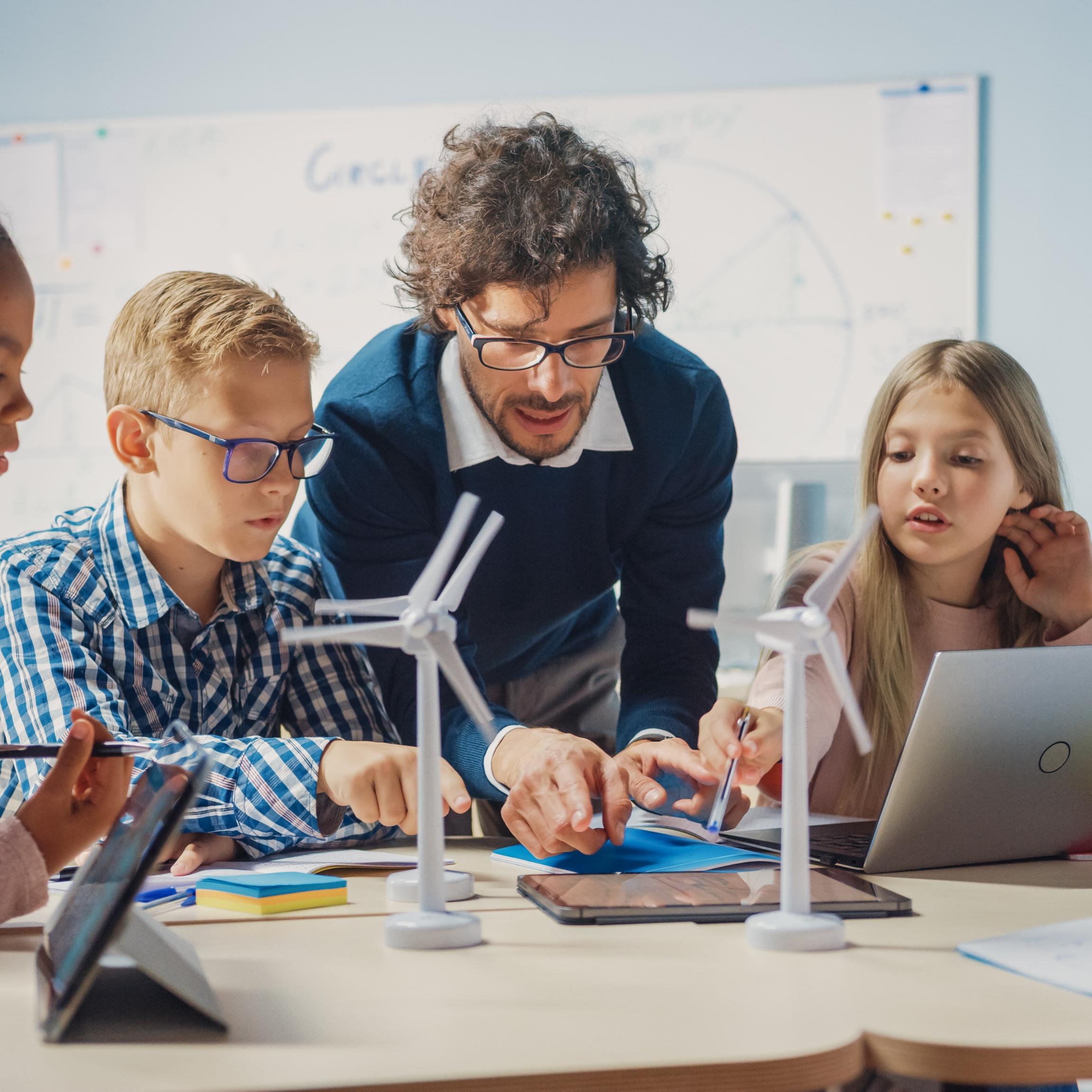 Man sieht Schüler*innen an einem Tisch mit Tablet, Laptop und Miniaturwindrädern. Ein Lehrer beugt sich zur Gruppe, um etwas zu erklären