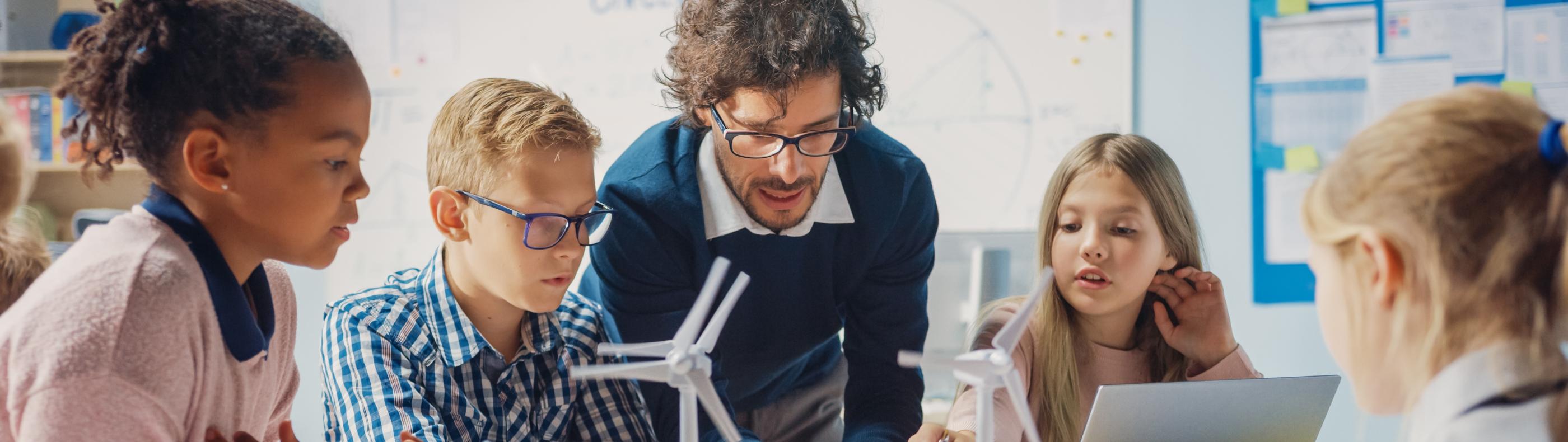 Man sieht Schüler*innen an einem Tisch mit Tablet, Laptop und Miniaturwindrädern. Ein Lehrer beugt sich zur Gruppe, um etwas zu erklären