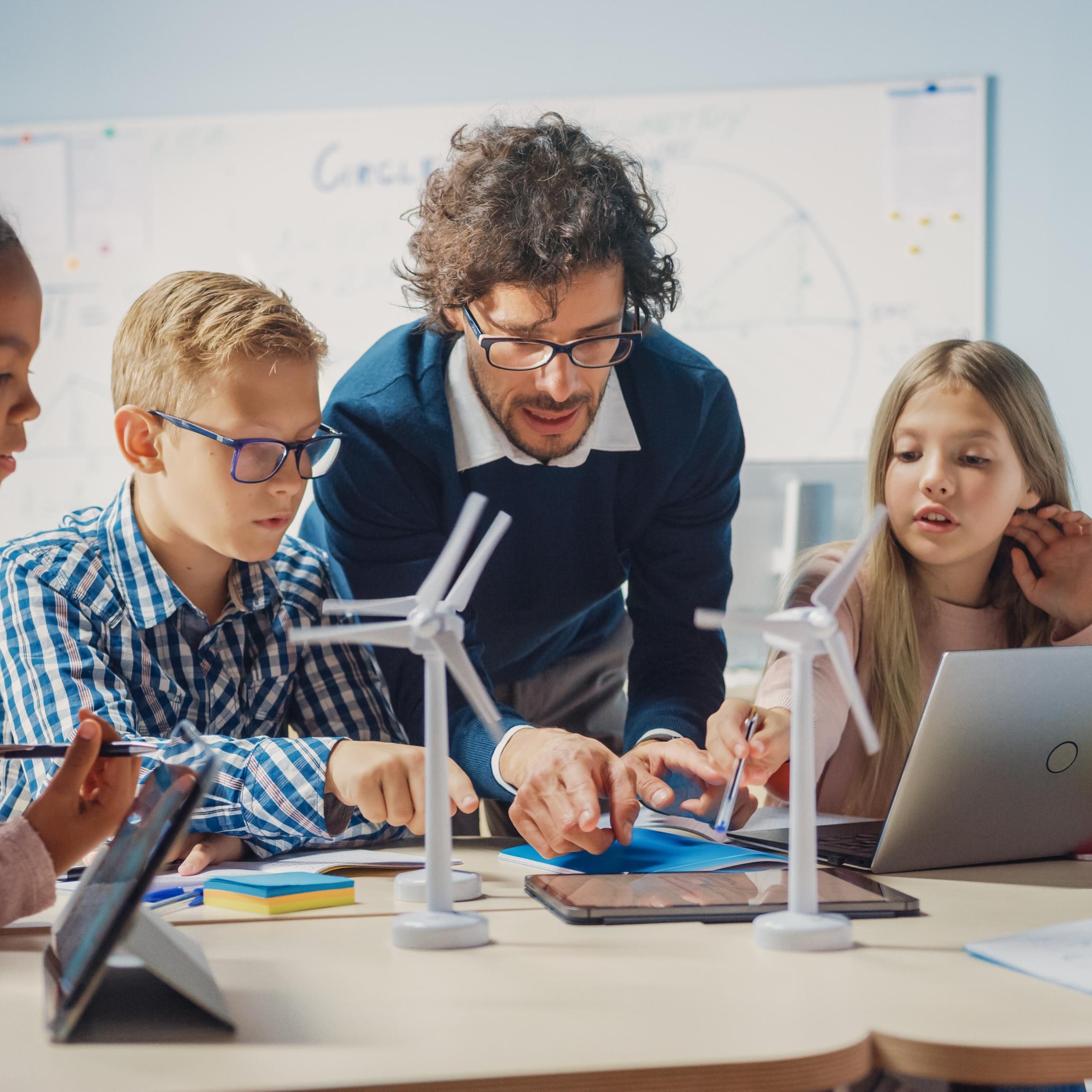 Man sieht Schüler*innen an einem Tisch mit Tablet, Laptop und Miniaturwindrädern. Ein Lehrer beugt sich zur Gruppe, um etwas zu erklären