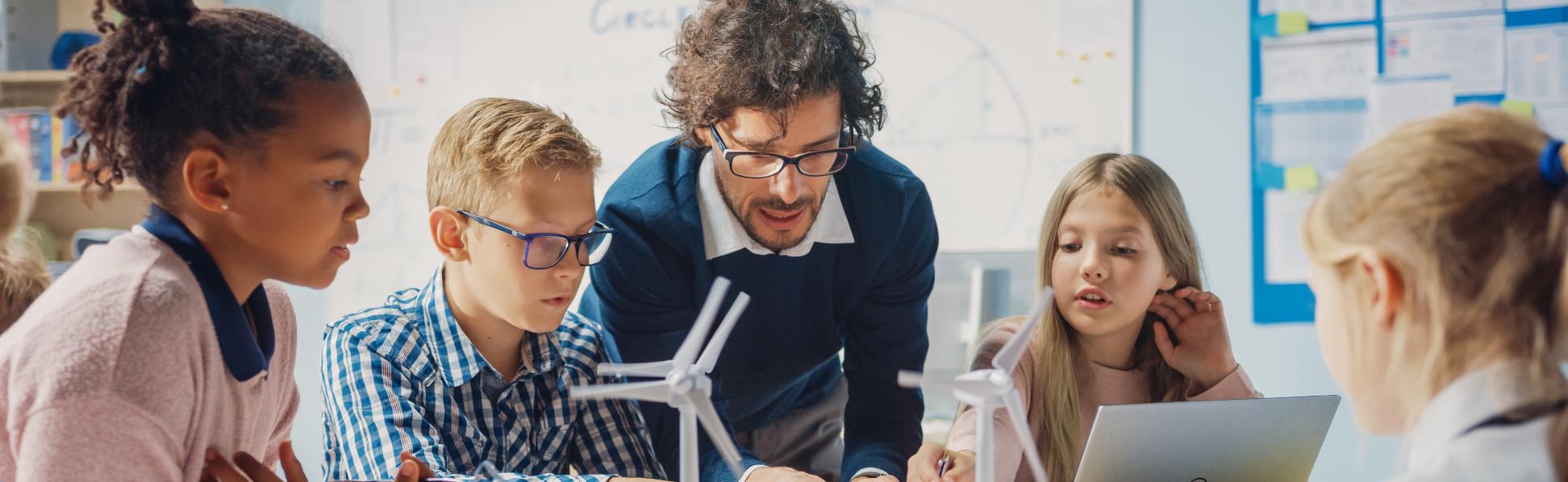 Man sieht Schüler*innen an einem Tisch mit Tablet, Laptop und Miniaturwindrädern. Ein Lehrer beugt sich zur Gruppe, um etwas zu erklären