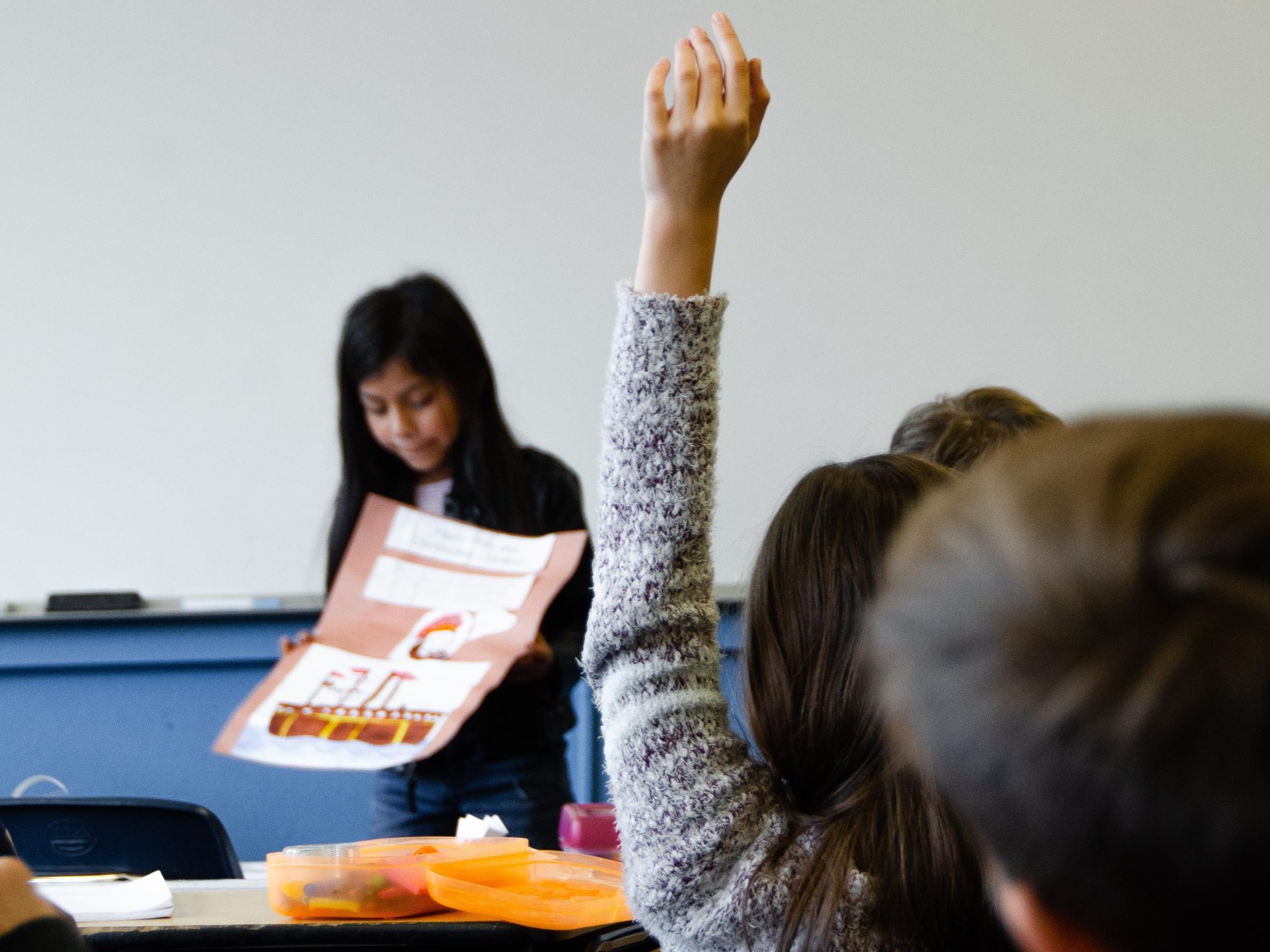 Man sieht einen Ausschnitt aus einem Klassenraum mit Schülern von hinten, von denen sich eine Schülerin meldet. Vorne steht ein Mädchen, das ein Plakat in den Händen hält.