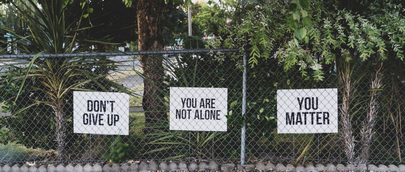 Man sieht einen Maschendrahtzaun, dahinter drei Schilder in Englisch beschriftet. Übersetzt: Geben Sie nicht auf! Sie sind nicht allein! Sie sind wichtig!