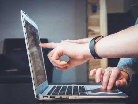 Man sieht einen aufgeklappten Laptop von der Seite. Eine Hand zeigt auf den Monitor, um etwas zu erklären