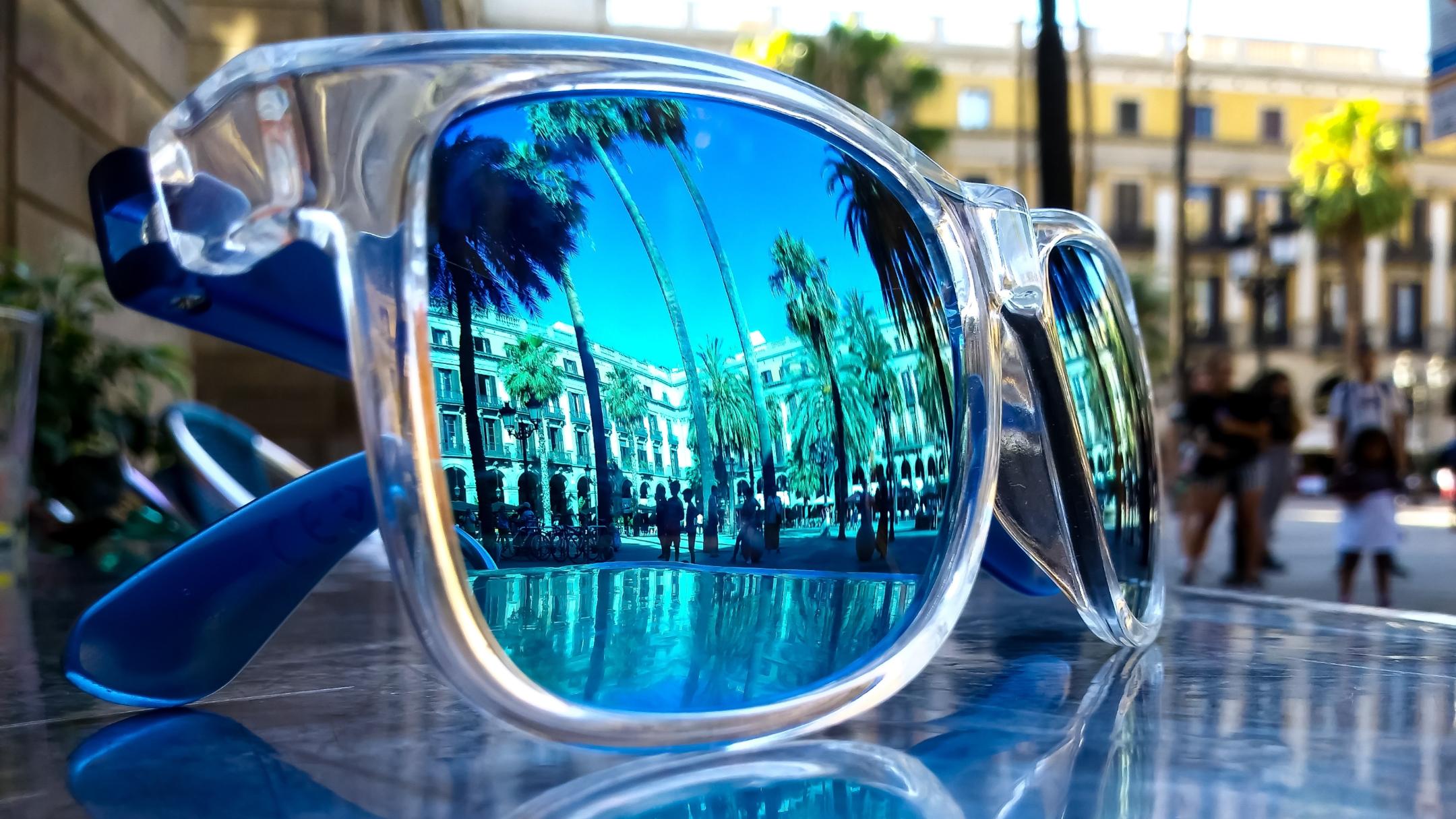 Eine Sonnenbrille liegt draußen auf einem Glastisch. In ihr spiegeln sich Palmen, Häuser und Menschen bei blauem Himmel.