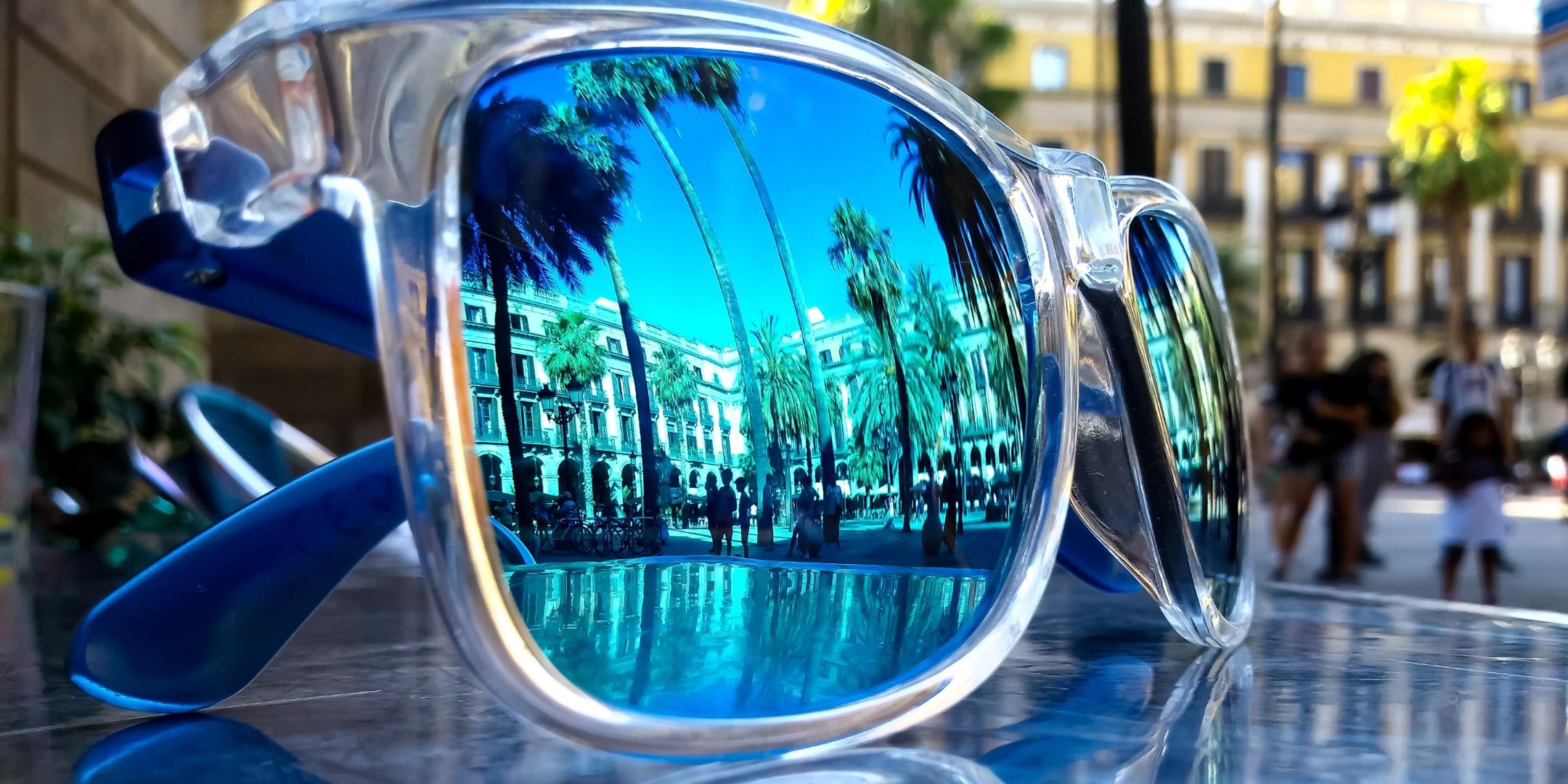 Eine Sonnenbrille liegt draußen auf einem Glastisch. In ihr spiegeln sich Palmen, Häuser und Menschen bei blauem Himmel.
