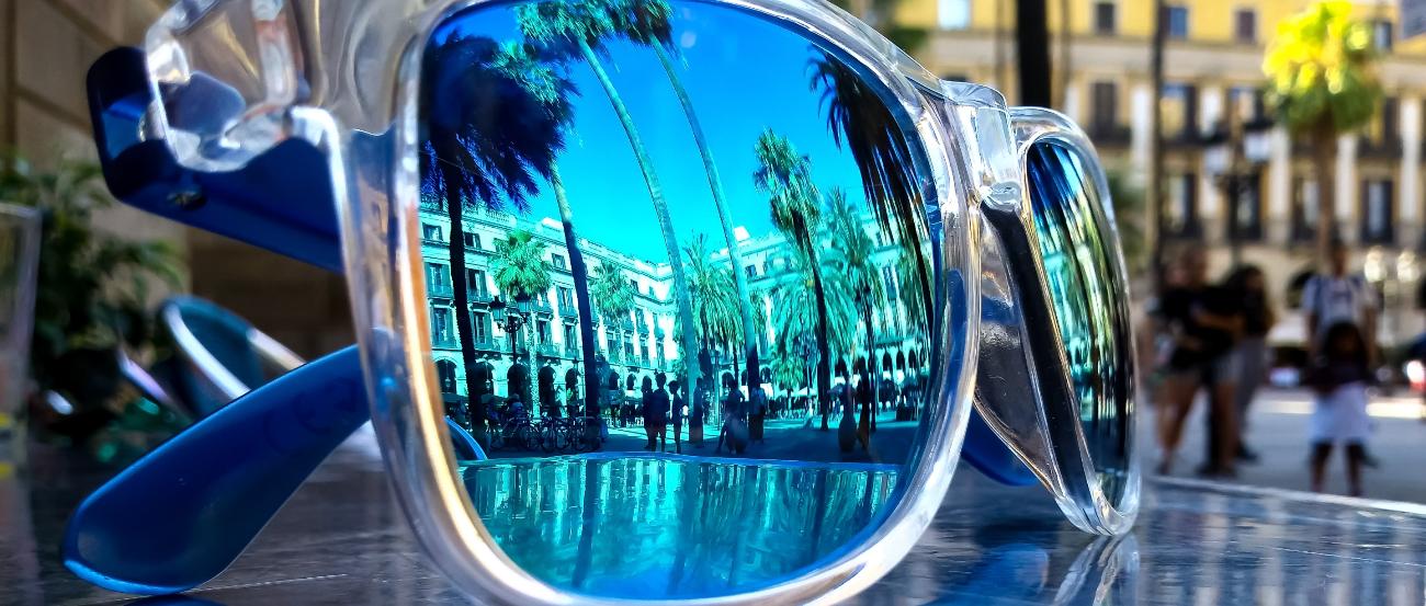 Eine Sonnenbrille liegt draußen auf einem Glastisch. In ihr spiegeln sich Palmen, Häuser und Menschen bei blauem Himmel.