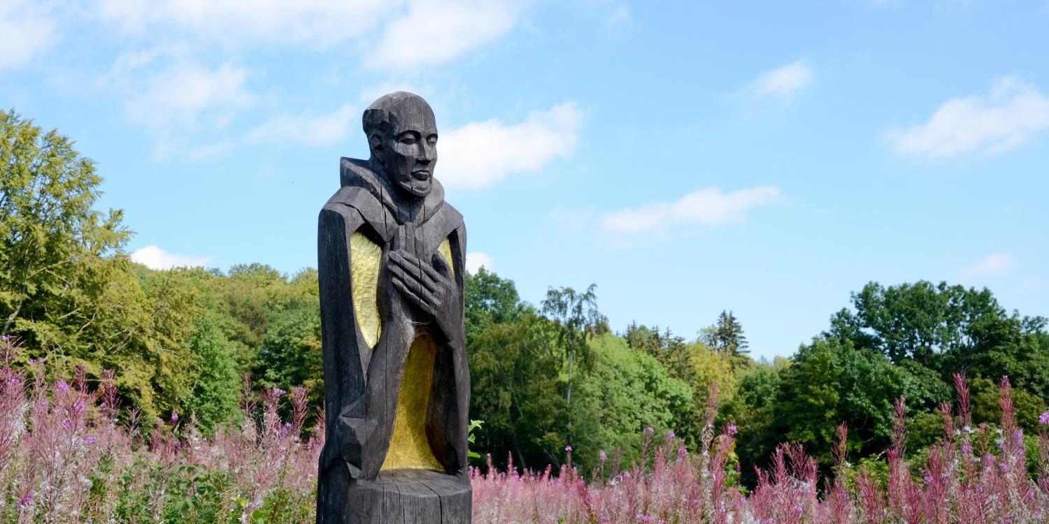 Holzskulptur im Park, umgeben von blühenden rosa Blumen und grünem Laub.