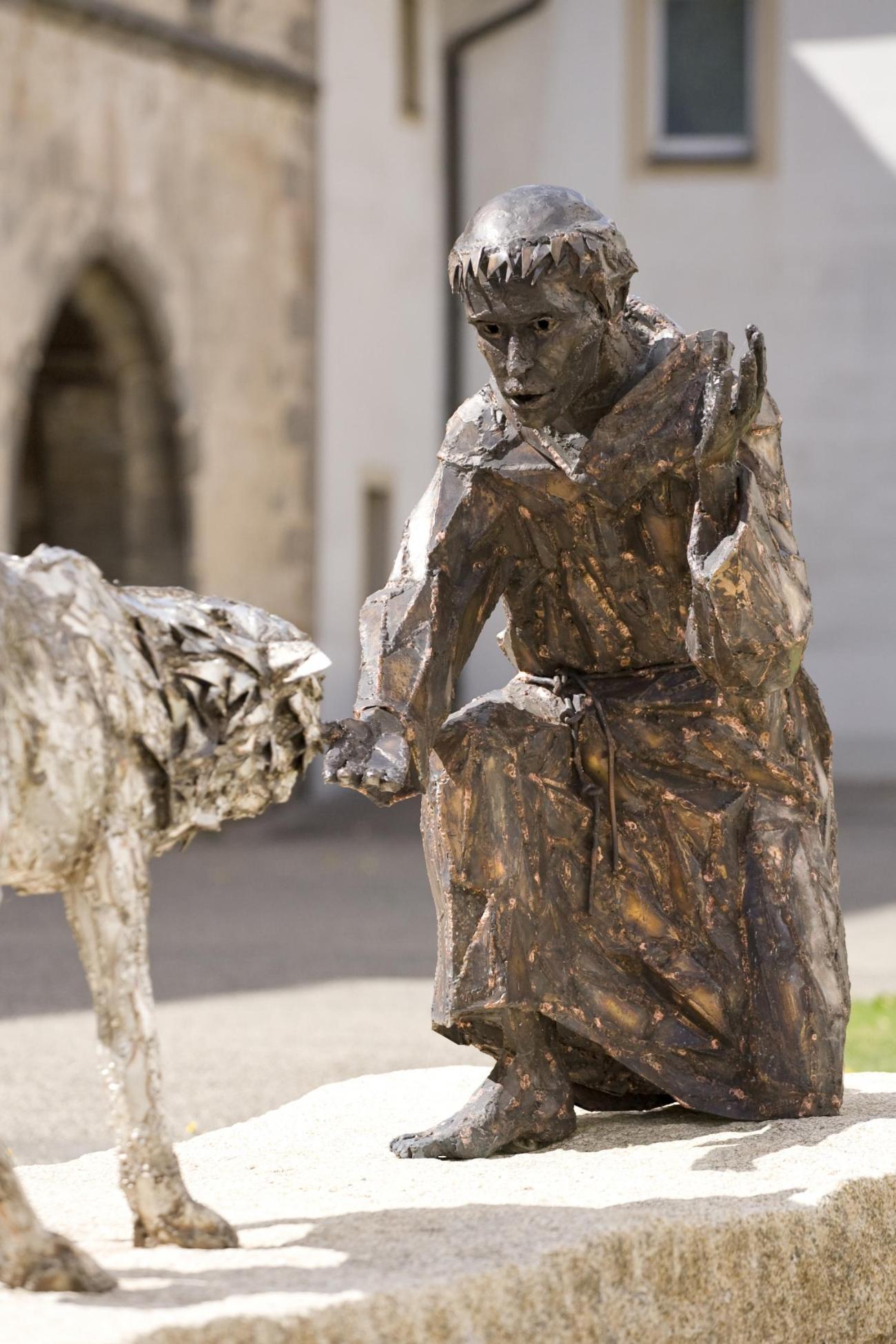 Bronzestatue eines knienden Mannes, der einem Wolf die Hand reicht.