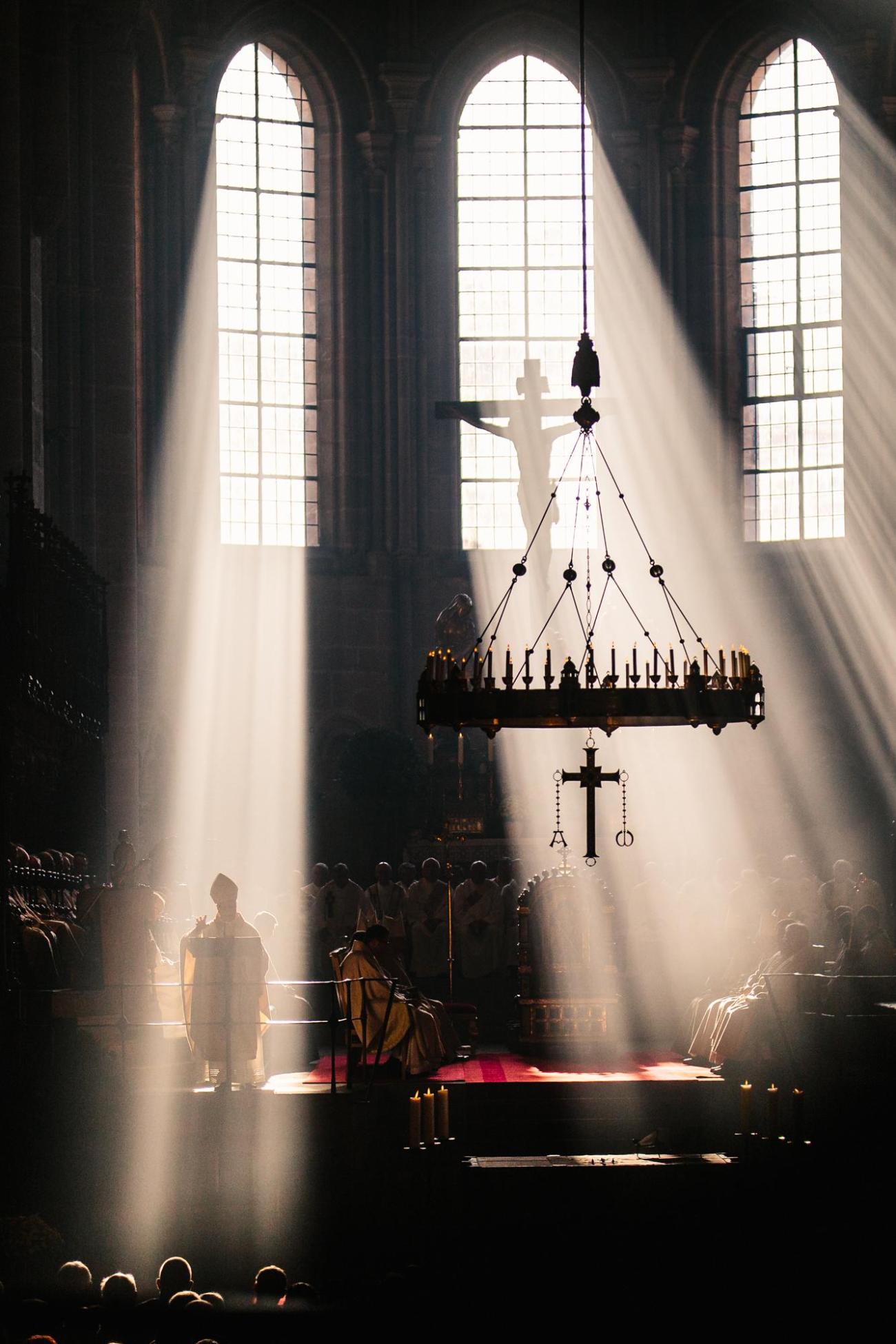 Sonnenstrahlen fallen durch große Fenster in den Bamberger Dom, links im Bild predigt ein Bischof am Ambo.