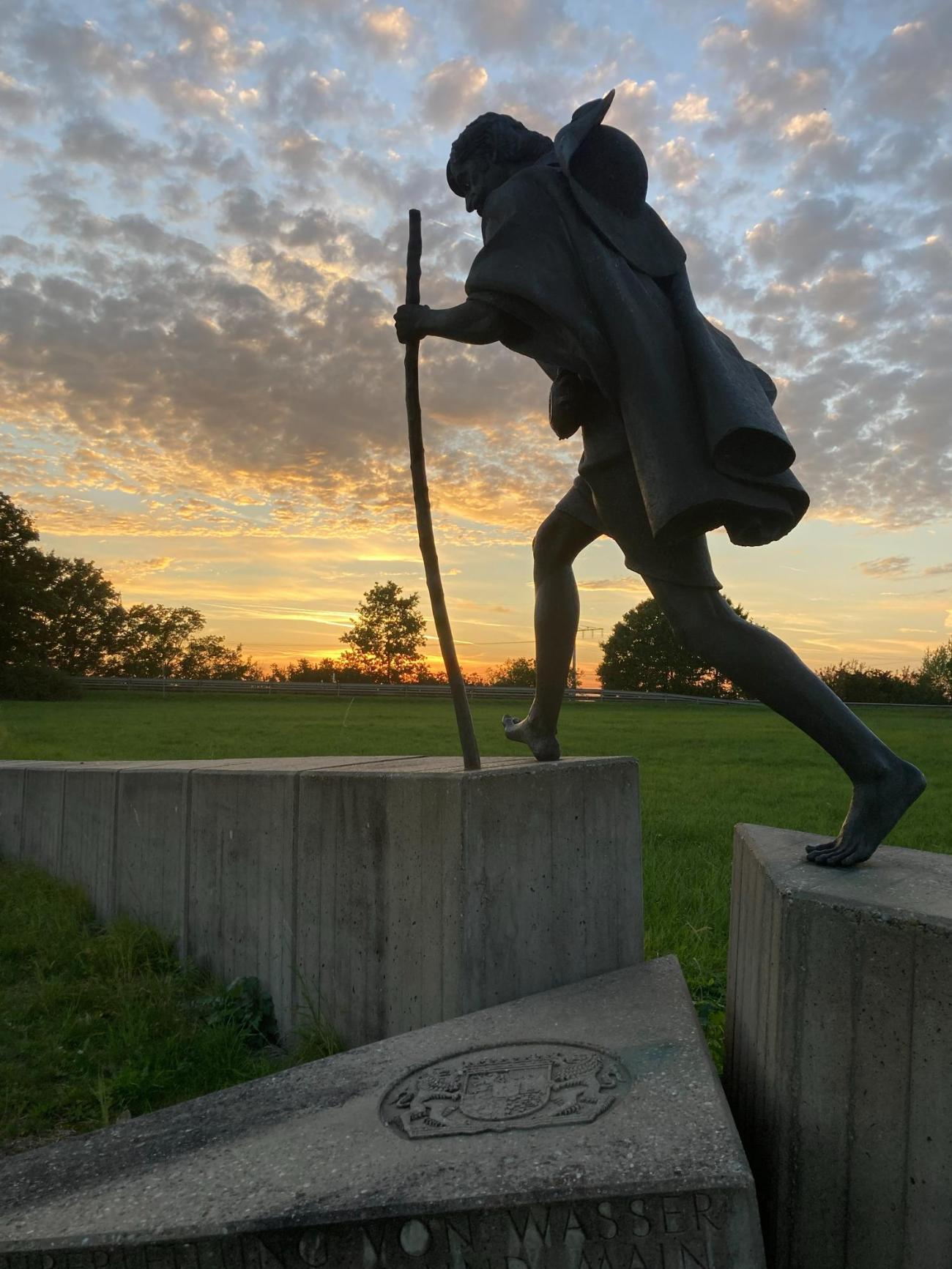 Bronzestatue eines gehenden Mannes mit Stock bei Sonnenuntergang auf Wiese.