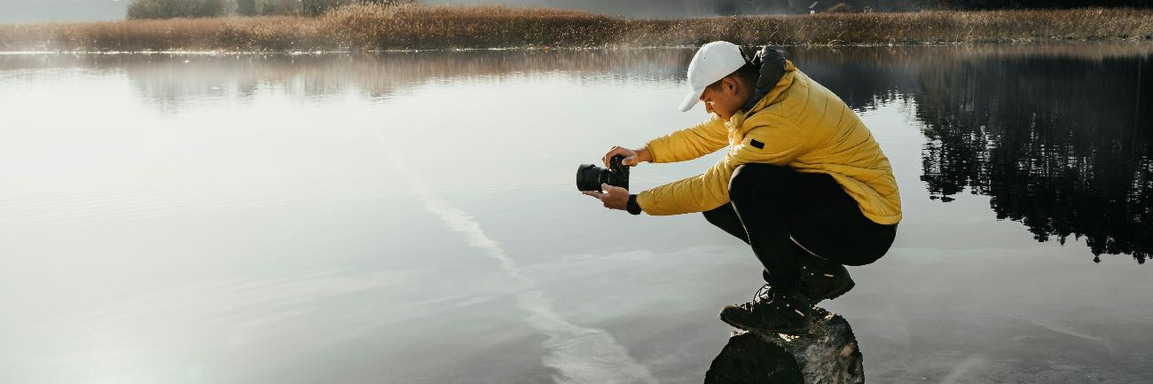 Ein Mann hockt auf einem Felsen und fotografiert einen See.