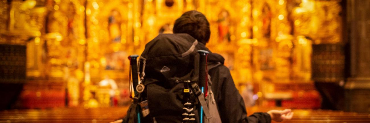 Pilger mit Rucksack, der dem kunstvollen Kirchenaltar zugewandt ist.