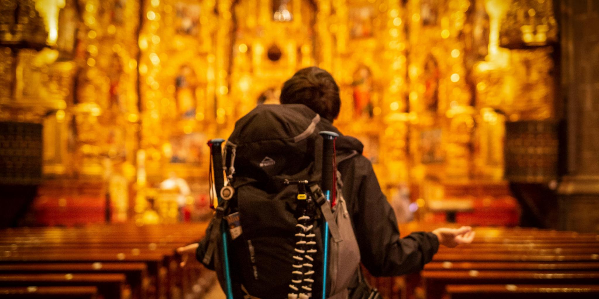 Pilger mit Rucksack, der dem kunstvollen Kirchenaltar zugewandt ist.