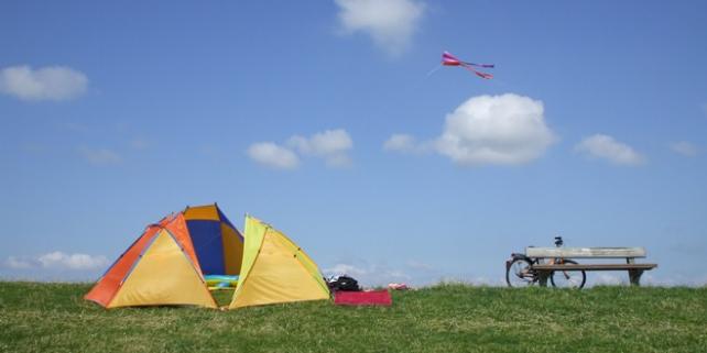 Drei Muschelzelte, eine Ruhebank, ein Fahrrad und ein kleiner Drache auf einem Deich unter blauem Himmel mit kleinen weißen Wolken