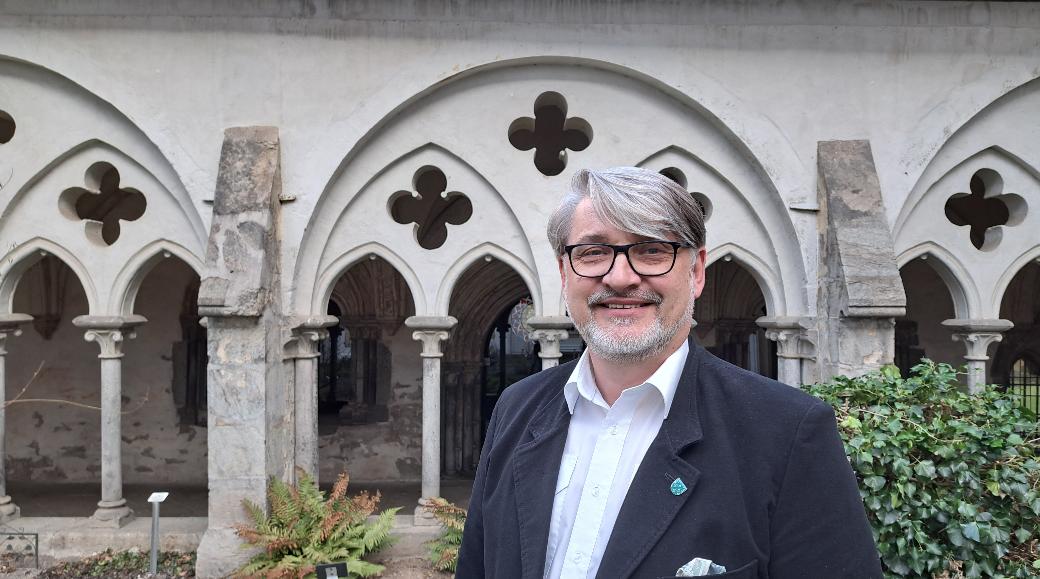 Ein Mann mittleren Alters mit grauen Haaren und Bart sowie Brille steht vor einem Kreuzgang.