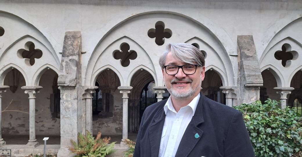 Ein Mann mittleren Alters mit grauen Haaren und Bart sowie Brille steht vor einem Kreuzgang.