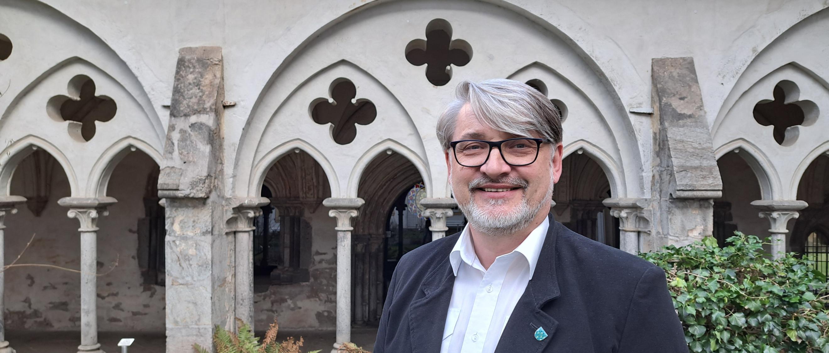 Ein Mann mittleren Alters mit grauen Haaren und Bart sowie Brille steht vor einem Kreuzgang.
