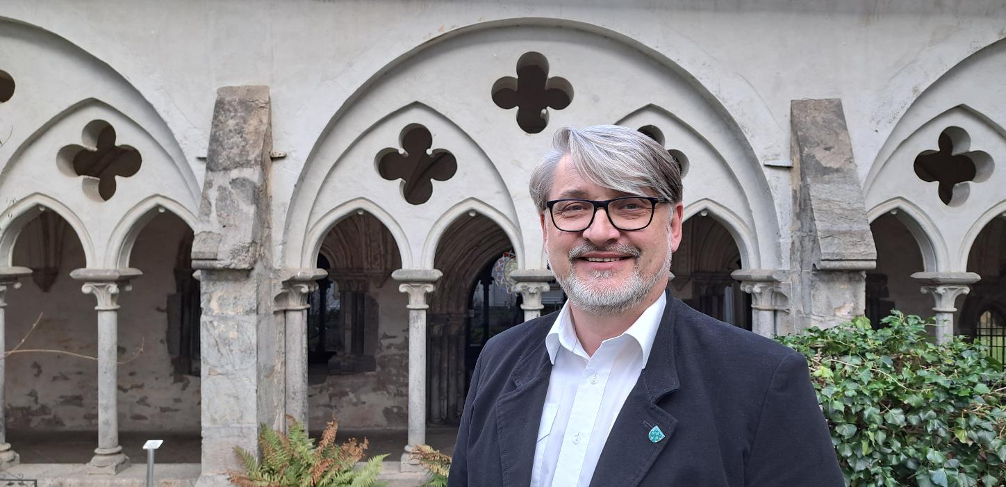 Ein Mann mittleren Alters mit grauen Haaren und Bart sowie Brille steht vor einem Kreuzgang.