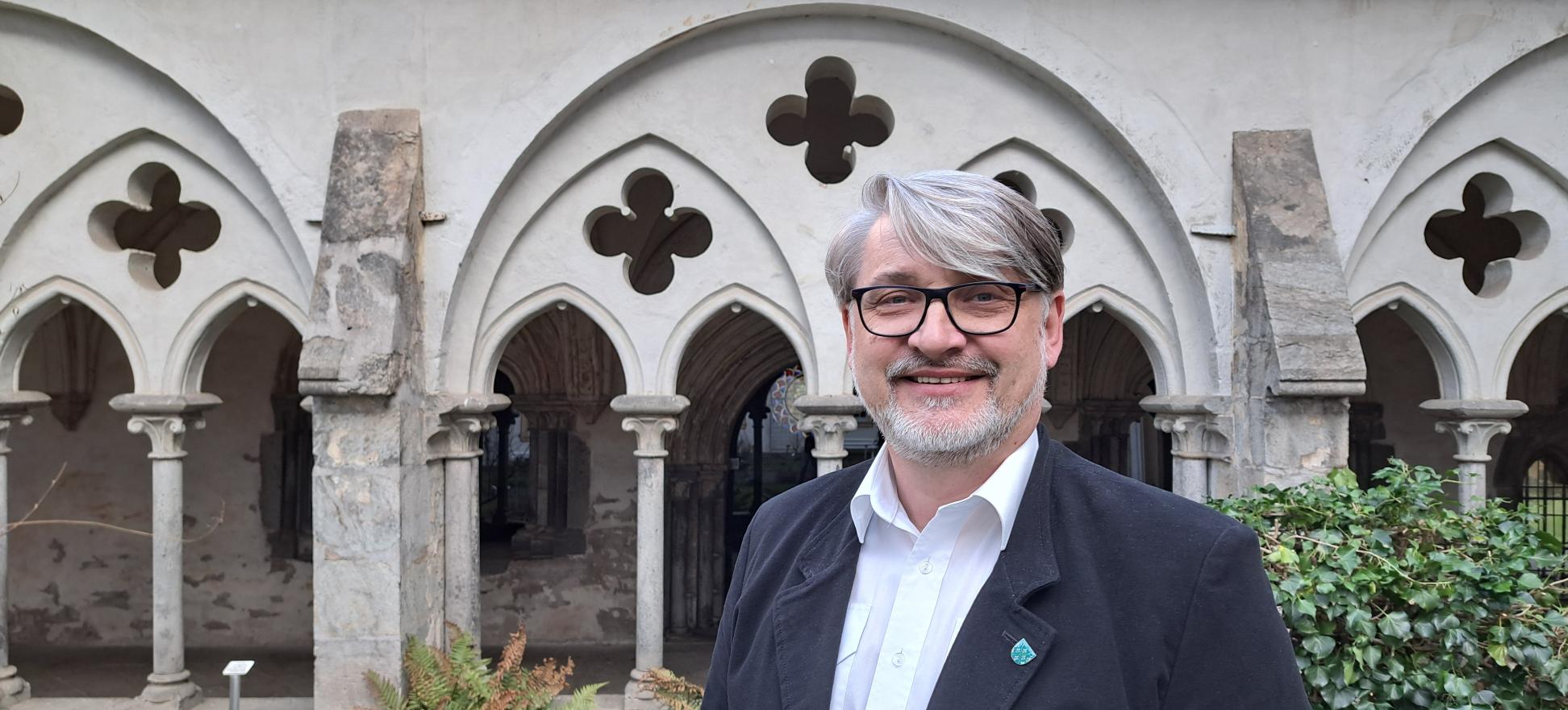 Ein Mann mittleren Alters mit grauen Haaren und Bart sowie Brille steht vor einem Kreuzgang.