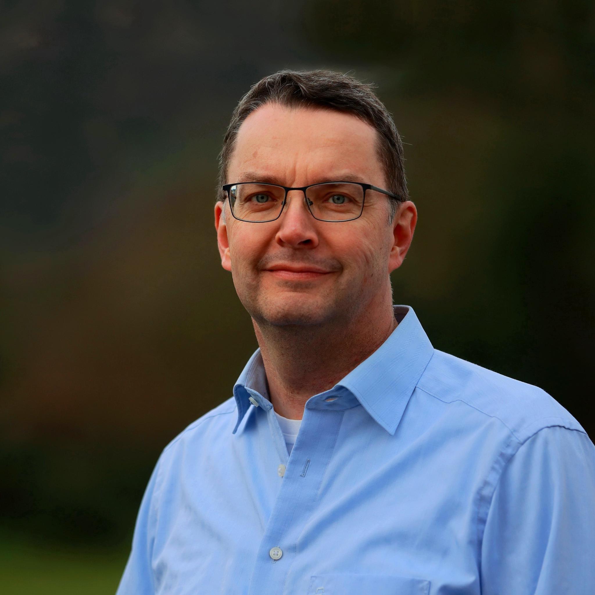 Man sieht ein Portrait eines Mannes mit dunklen Haaren und einer Brille, der leicht in die Kamera lächelt