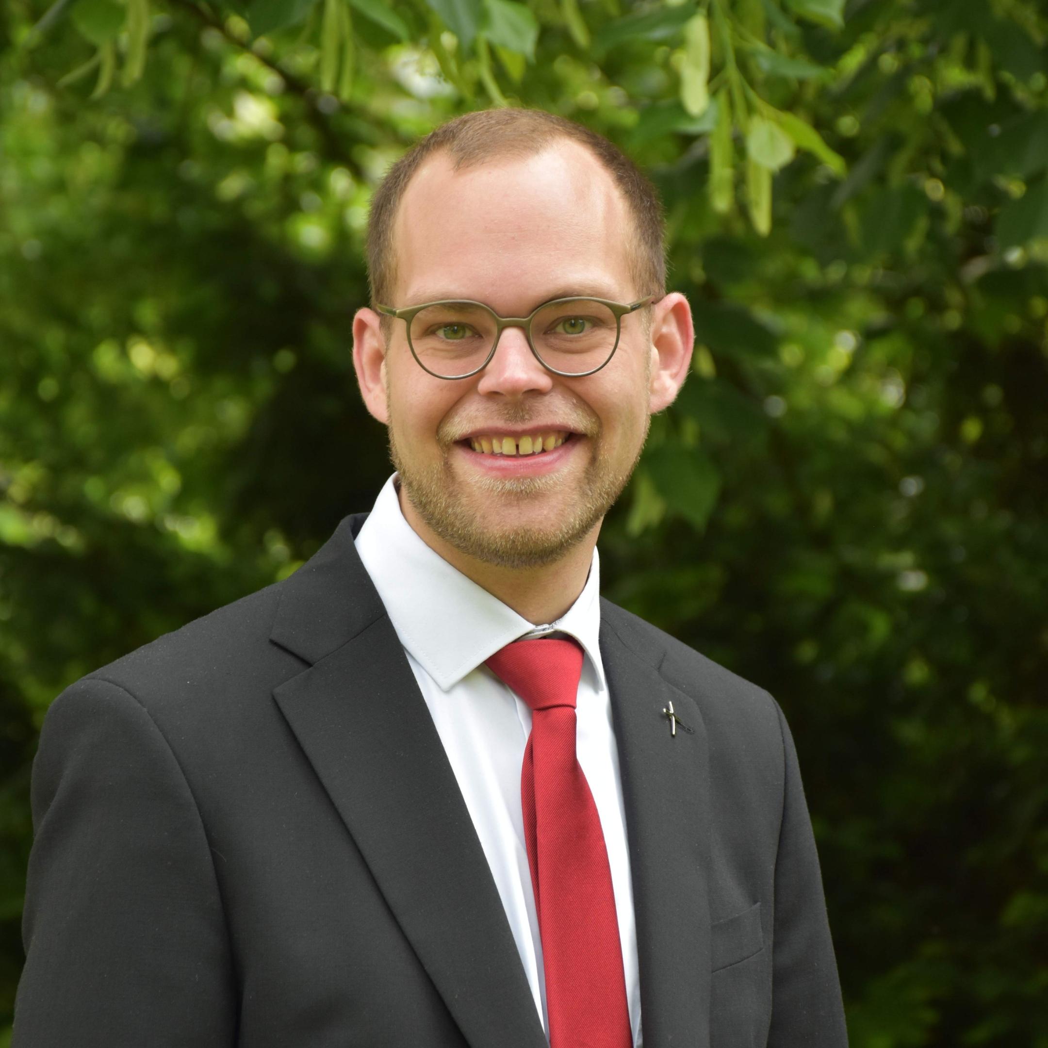 Man sieht Mathias Kremer Gaub im Anzug und Krawatte. Er trägt einen Bart, Brille und lächelt in die Kamera.