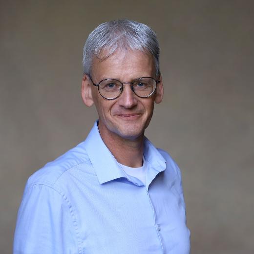Ein Mann im blauen Hemd, Brille und grauen Haaren lächelt in die Kamera