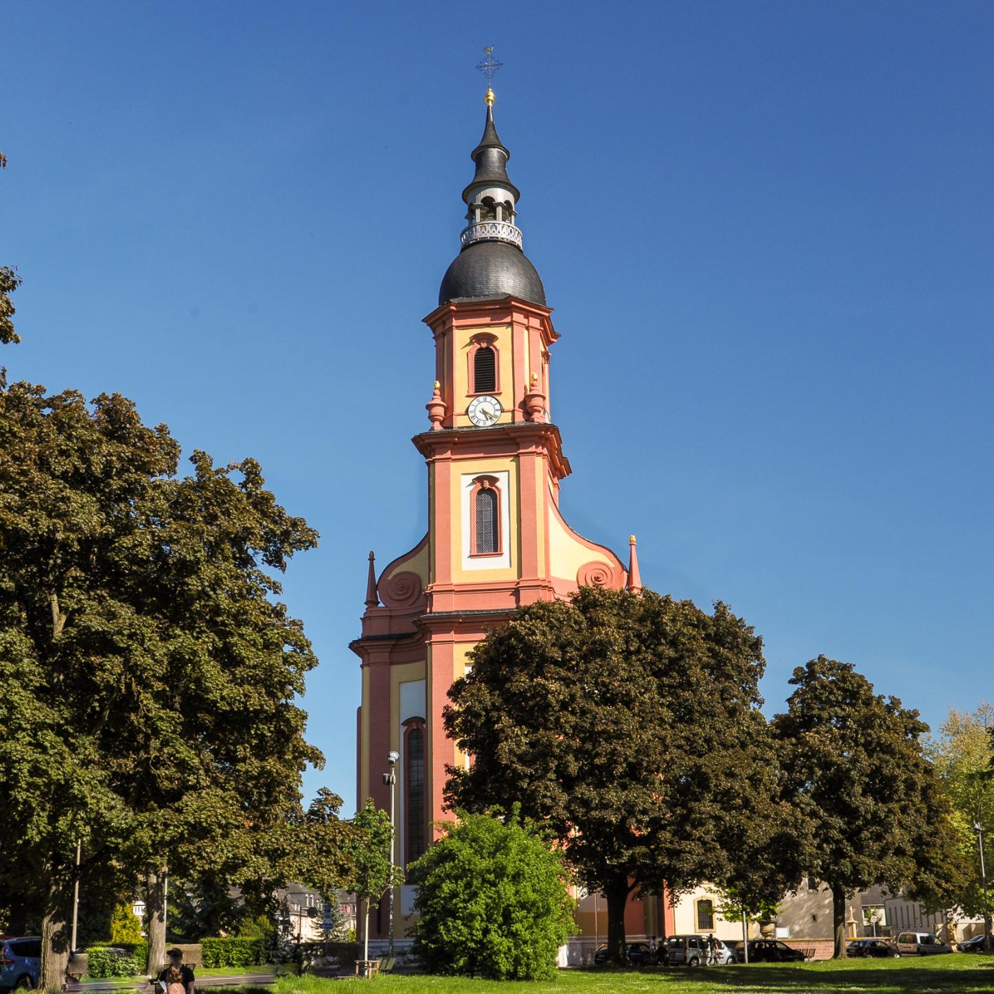 Kirche mit hohem Turm in Rot-Gelb-Tönen, umgeben von Bäumen