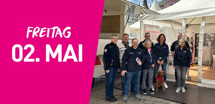 Eine Gruppe der Malteser steht vor einem Infozelt. Links im Bild steht auf pinkfarbenem Hintergrund Freitag zweiter Mai