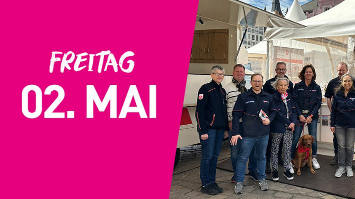 Eine Gruppe der Malteser steht vor einem Infozelt. Links im Bild steht auf pinkfarbenem Hintergrund Freitag zweiter Mai