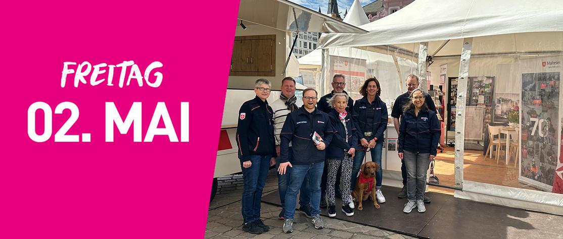 Eine Gruppe der Malteser steht vor einem Infozelt. Links im Bild steht auf pinkfarbenem Hintergrund Freitag zweiter Mai