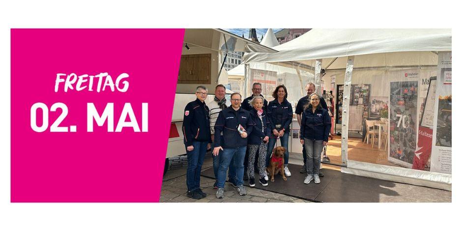 Eine Gruppe der Malteser steht vor einem Infozelt. Links im Bild steht auf pinkfarbenem Hintergrund Freitag zweiter Mai