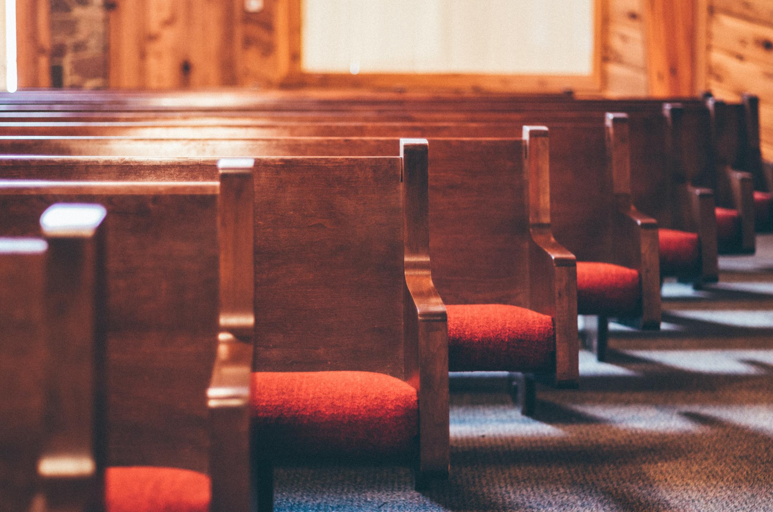 Das Bild zeigt eine Reihe mir dunkelbraunen Kirchenbänken mit roten Sitzpolstern