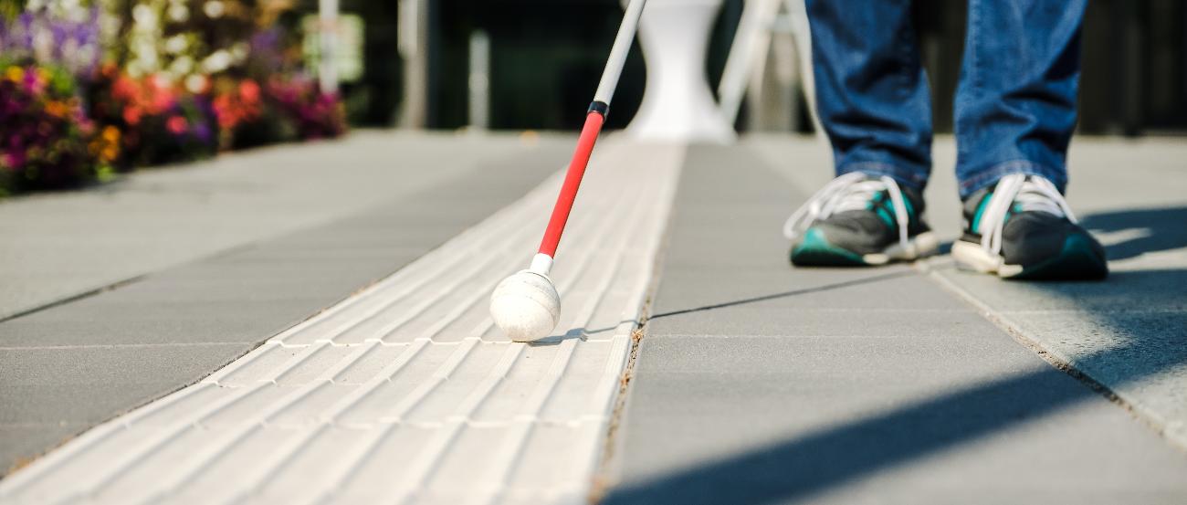 Man sieht bodennah einen Blindenstock, der einen Gehsteig betastet